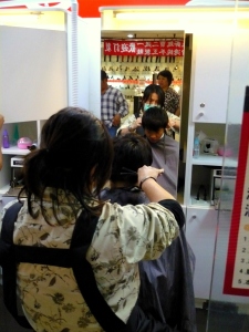 Barber, Sam Zhu is skillfully at work with one of his clientes, as others [behind, left] are coming in queue to purchase their tickets.  